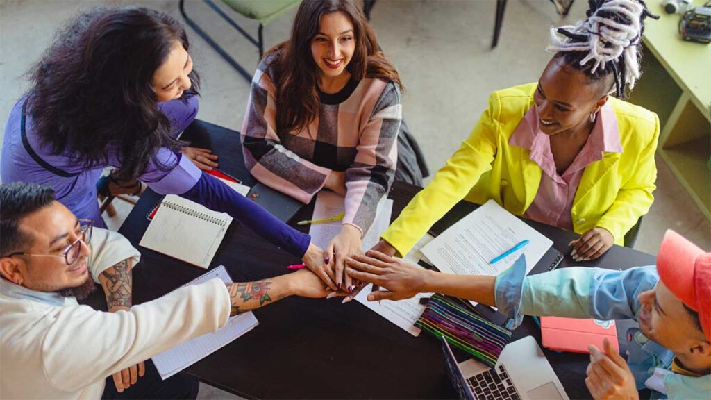 A group of five diverse coworkers cheerfully joining hands in a show of teamwork around a table